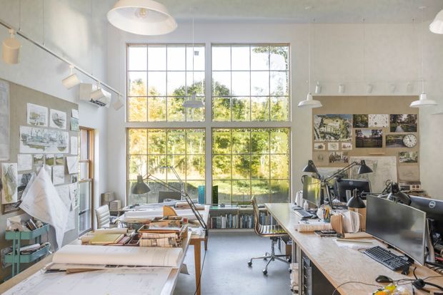 Bright, spacious architectural design studio with large industrial-style grid windows, drafting tables, computers, task lamps, bookshelves, and mood boards. The workspace has pale walls, exposed utilitarian finishes, abundant natural light, and an organised professional atmosphere.