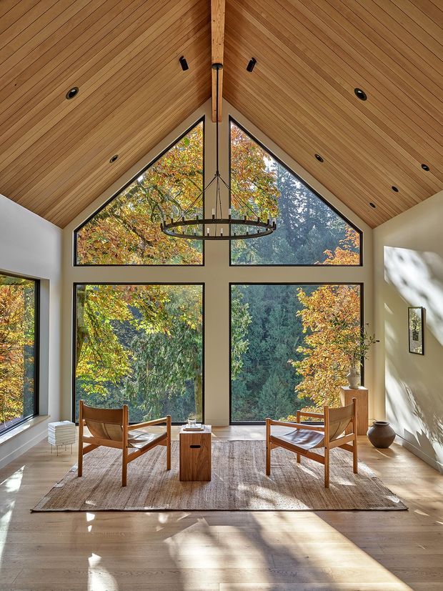 A serene, double-height sitting room with a dramatic vaulted timber-lined ceiling and exposed central ridge beam. Large black-framed triangular and rectangular windows form a geometric glazed wall overlooking dense autumn woodland. Two mid-century-inspired timber lounge chairs face each other around a small wood side table on a neutral woven rug, with pale timber flooring, warm white walls, recessed ceiling lights, and a sculptural circular pendant. Sunlight creates strong dappled shadows across the floor.