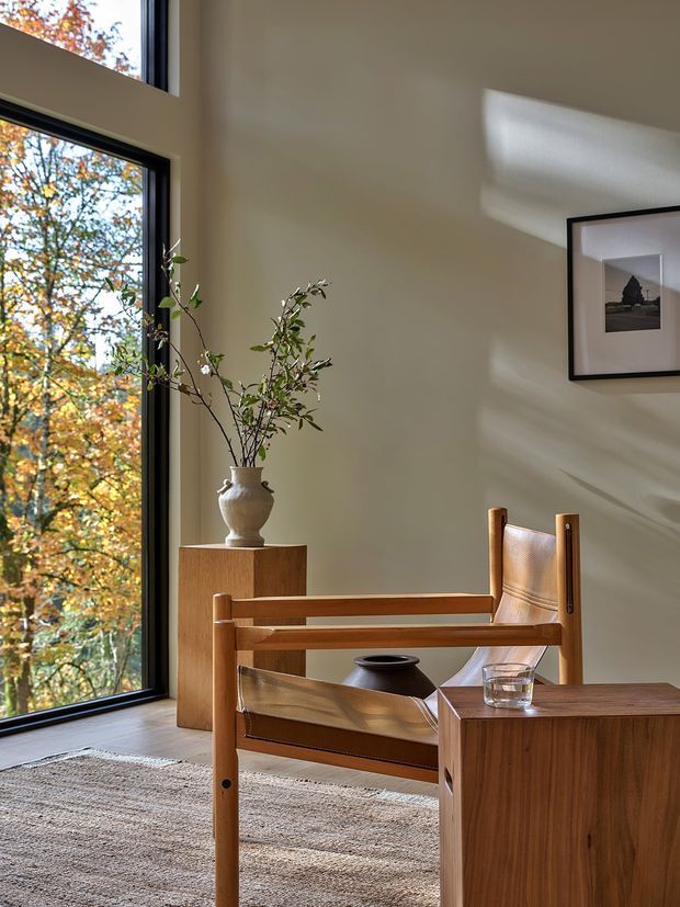 A warmly lit, minimalist sitting area with a large black-framed window overlooking autumn trees. The room features a sculptural wooden armchair with tan leather upholstery, a matching wood side cabinet, a textured neutral rug, a ceramic vase with branches, and framed artwork. Strong sunlight creates soft geometric shadows across the beige wall.