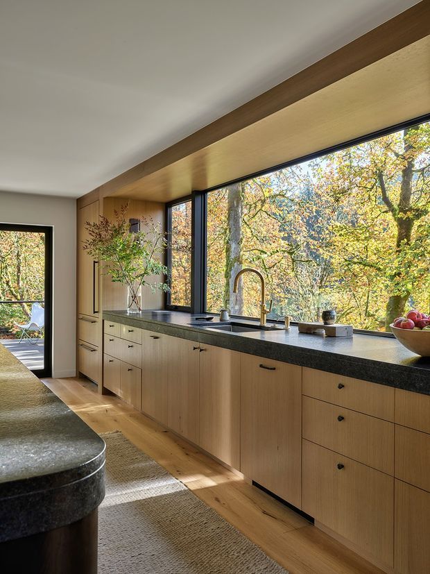 Warm contemporary kitchen with a long wall of light timber cabinetry, dark stone benchtops, black-framed floor-to-ceiling windows, and a timber-lined ceiling recess. The kitchen opens toward a glazed exterior doorway and overlooks leafy autumn vegetation. A rounded dark stone island is partially visible in the foreground.