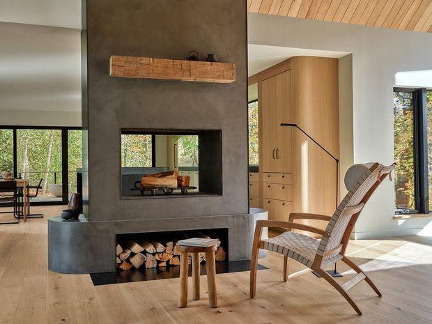 Warm, light-filled contemporary living room centered on a tall dark plaster fireplace wall with an inset firebox, timber mantel shelf, and neatly stored firewood. Pale timber flooring and a slatted timber ceiling complement built-in timber cabinetry, large windows, a woven lounge chair, and a small timber stool. Forested views are visible through the glazing.