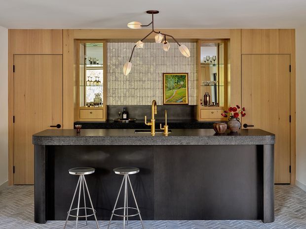 A refined kitchen with a large dark stone island and integrated seating, framed by warm timber cabinetry and concealed full-height pantry doors. The rear wall features a textured grey tiled splashback, open display shelving with glassware, a dark benchtop, and brass-toned tapware. A sculptural multi-arm pendant light hangs above the island, while a herringbone-patterned grey floor adds texture and visual interest.