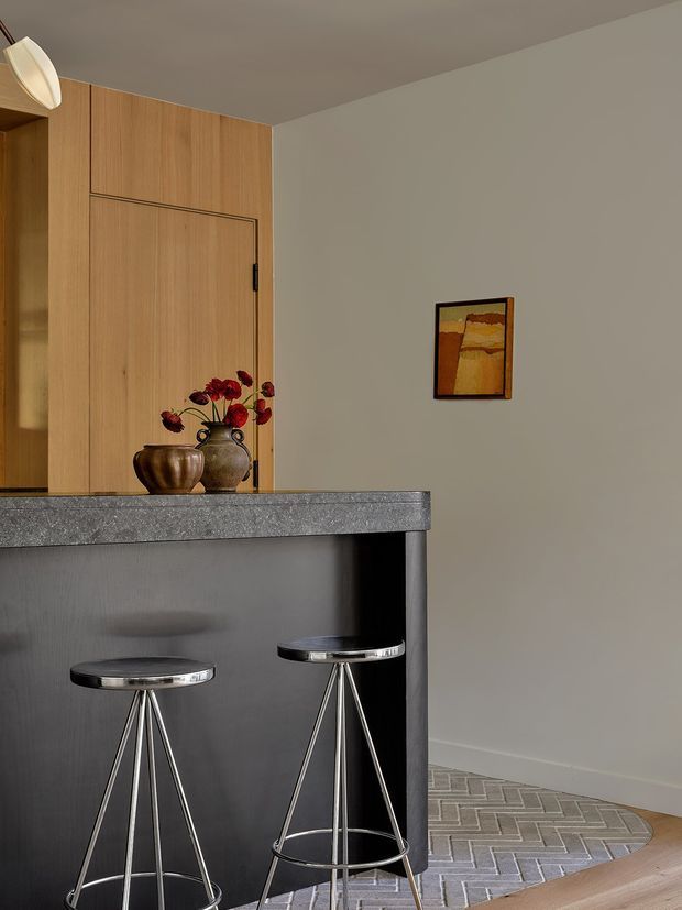 A refined contemporary kitchen and breakfast bar with warm timber full-height cabinetry, a dark charcoal island with a textured grey stone benchtop, and two slim chrome bar stools. A ceramic vase arrangement and small abstract artwork add understated character. The room has a neutral palette, soft lighting, and a patterned grey tiled floor transitioning to timber flooring.