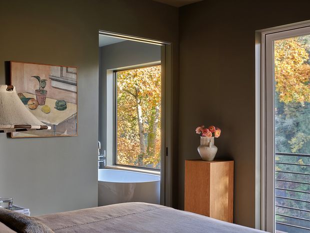 A warm, moody bedroom interior viewed toward an adjoining bathroom and a large window. The room has dark taupe painted walls, a low-profile bed with a neutral textured cover, a timber pedestal cabinet with a ceramic vase, and framed artwork. A freestanding or partially visible bathtub is seen through the open doorway, while autumn trees are visible outside through the glazing.