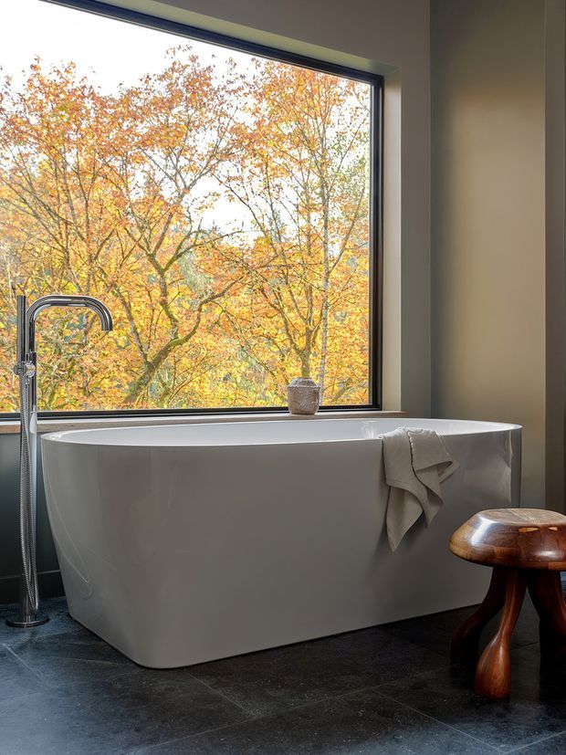 A serene, contemporary bathroom featuring a freestanding white bathtub positioned beneath a large black-framed picture window overlooking autumn trees. A tall floor-mounted chrome bath filler stands beside the tub, with a folded neutral towel draped over the rim and a sculptural dark wooden stool nearby. The room has dark stone-look floor tiles, warm neutral walls, and a strong connection to the surrounding landscape.