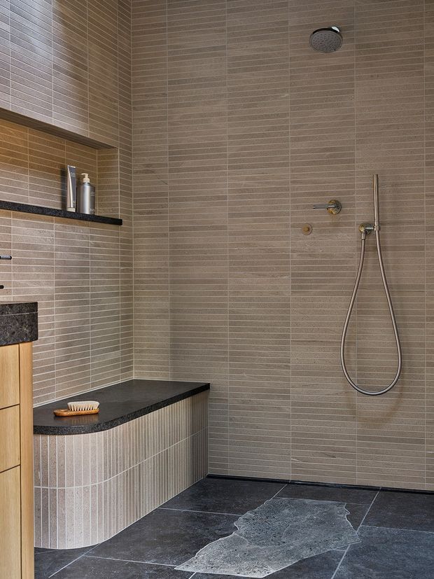 Contemporary walk-in shower room with warm taupe linear wall tiles, dark charcoal stone flooring, and a curved built-in bench with vertical fluted tile detailing. The space includes a rainfall showerhead, handheld shower, recessed shelf, and dark stone ledge, creating a refined spa-like bathroom interior.