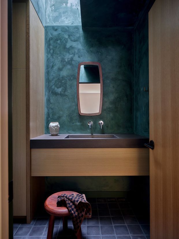 A compact, moody powder room viewed through an open timber door. The room features a floating timber vanity with a dark stone benchtop and integrated rectangular basin, wall-mounted chrome taps, a mottled deep green plaster feature wall, a sculptural framed mirror, dark tiled flooring, and a small upholstered stool. Tall timber cabinetry or wall panelling frames the vanity.