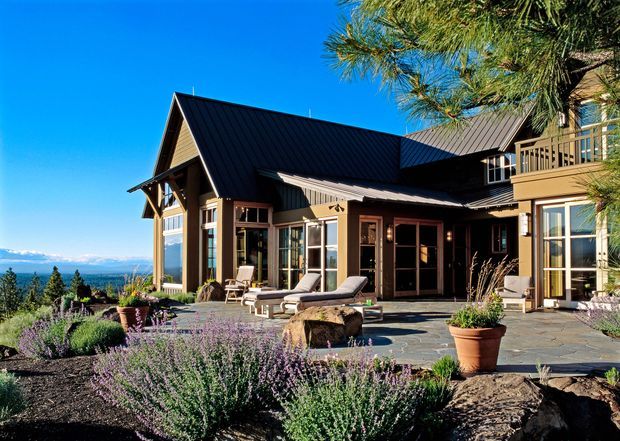 Exterior view of a substantial multi-level home set on a landscaped, rocky site. The building has dark standing-seam pitched roofs, timber-framed gabled sections, extensive glazing, and a broad stone-paved terrace with outdoor seating. Low flowering shrubs, large rocks, and potted plants surround the terrace, while a distant mountainous landscape is visible beyond.