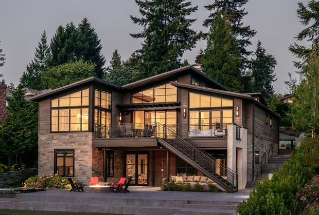 Exterior view of a large contemporary multi-level house set among mature evergreen trees. The home features broad floor-to-ceiling glazing with warm interior illumination, dark timber-toned cladding, stone-faced lower walls, prominent gabled roof forms, balconies, and an exterior stair leading to an elevated entry terrace. Landscaped planting and paved areas surround the facade.