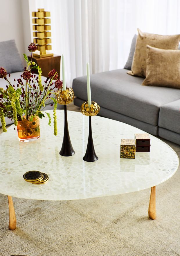 Bright, refined living room vignette centred on a large oval coffee table with a pale white textured mosaic-like surface and light timber legs. The table is styled with black-and-gold taper candleholders, a floral arrangement in an amber glass vase, small decorative boxes and a coaster. A large grey sectional sofa with plush tan cushions sits behind it, with sheer white curtains and a gold sculptural decorative object adding visual interest. The room has a warm, polished contemporary feel.