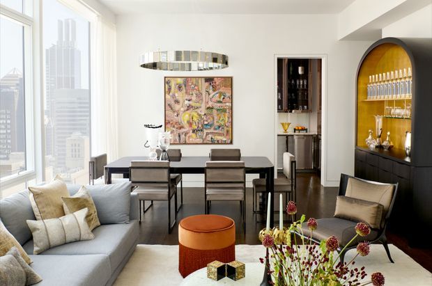 Bright, sophisticated open-plan living and dining area in a high-rise apartment, with floor-to-ceiling city views, a pale sectional sofa, dark dining table and chairs, contemporary artwork, statement pendant lighting, and a dramatic arched built-in bar with illuminated amber-toned backing and glassware.
