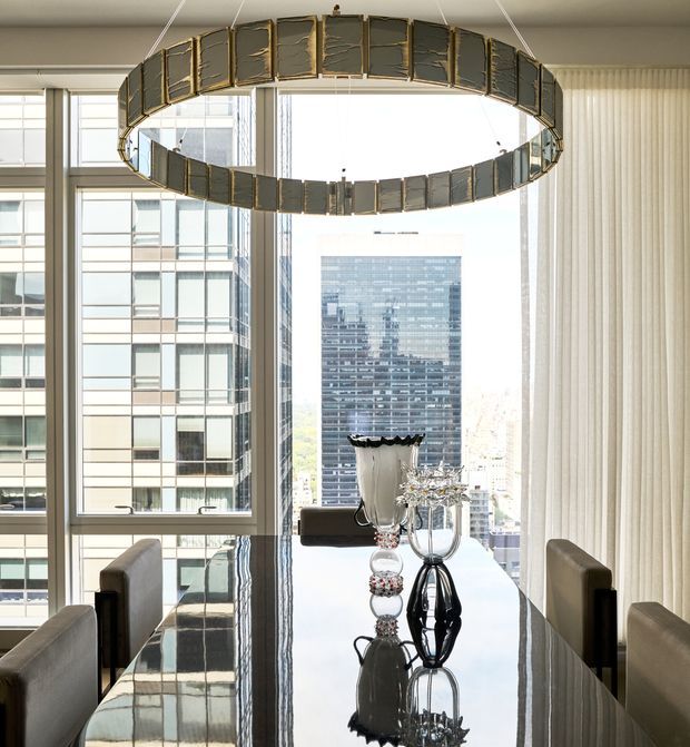 Bright high-rise dining room with a long glossy dark dining table, upholstered dining chairs, floor-to-ceiling windows overlooking an urban skyline, sheer vertical blinds, and a large suspended circular sculptural pendant light. Reflective decorative vessels form a centrepiece on the table.