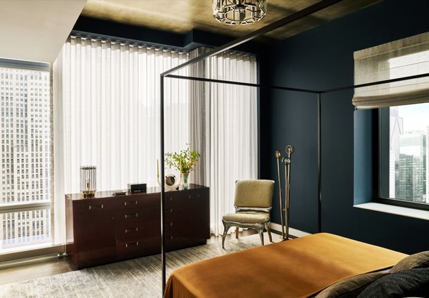 A sophisticated bedroom in a high-rise apartment, featuring deep navy walls, floor-to-ceiling sheer curtains, large city-view windows, a dark wood sideboard, an upholstered accent chair, and a bed dressed in warm ochre-brown bedding. A black-framed glass partition separates the sleeping area, while a sculptural ceiling light and textured ceiling add visual interest.