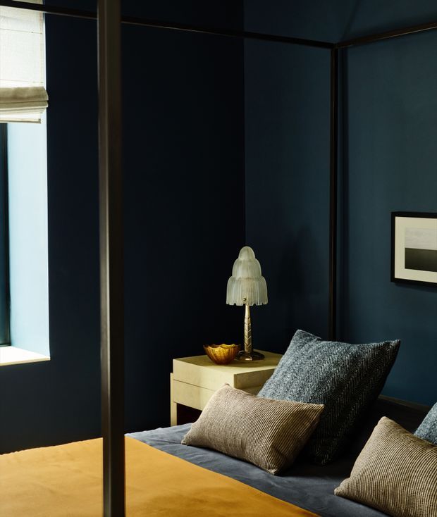 Moody, sophisticated bedroom corner with deep navy-blue walls, dark trim, and a partially visible bed layered with textured charcoal and taupe cushions. A pale wood bedside table holds a sculptural glass lamp and small decorative bowl. A window with a light Roman blind brings in a narrow band of daylight, while framed minimalist artwork adds a refined finishing touch.