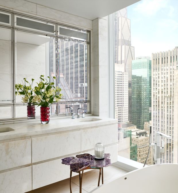 Bright, high-rise bathroom with expansive city views through floor-to-ceiling windows. The room features a pale marble-clad vanity with integrated basins and chrome tapware, framed windows, a freestanding white bathtub in the foreground, and decorative flower arrangements on the vanity. The space feels luxurious, airy, and contemporary.