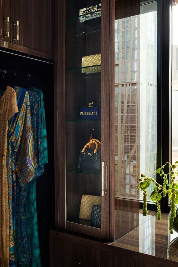 A refined walk-in wardrobe with dark timber built-in cabinetry, brass-toned hardware, glass-front display shelving, hanging patterned garments, handbags and accessories. A full-height window or glazed panel reflects a nearby high-rise building, while a polished dark surface and trailing plant add visual interest. The space has a rich, luxurious and moody character.