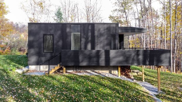 A compact modern dark-clad house set within a wooded, gently sloping site. The box-like dwelling has a flat roof, tall narrow windows, and a raised deck or terrace projecting over the landscape on slender supports. Autumn trees surround the structure, with a small stair and walkway leading to the entrance.