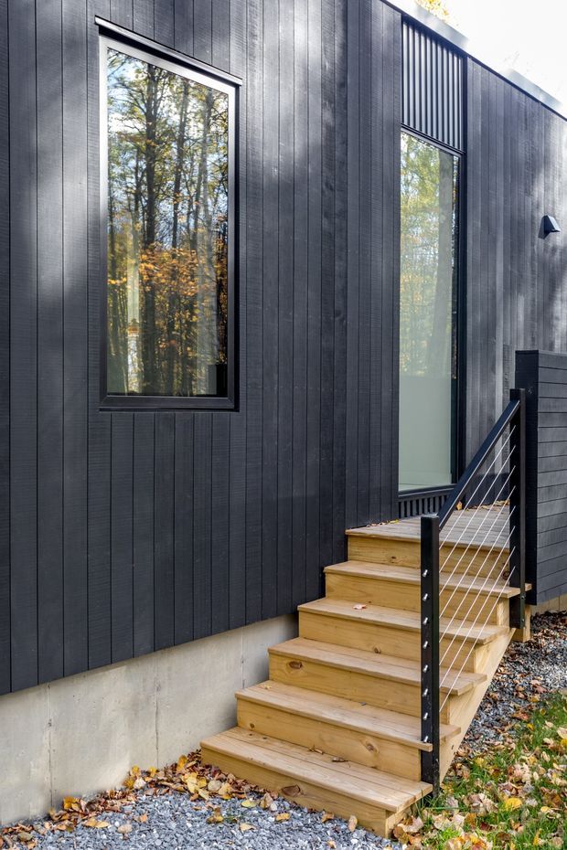 Exterior view of a compact modern building clad in dark vertical timber boards, with black-framed windows, a timber stair and simple cable railing leading to an entry door. The building is set beside a wooded landscape, with gravel and fallen leaves at ground level.