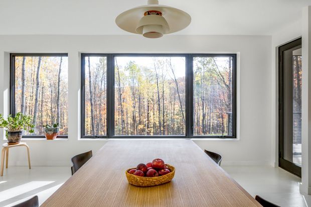 Bright minimalist dining room with a long light-wood dining table, dark dining chairs, large black-framed windows overlooking a wooded landscape, white walls and floor, potted plants, and a sculptural cream pendant light.