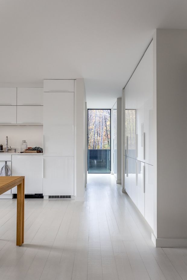 A bright, minimalist open-plan kitchen and dining area viewed along a narrow hallway. White flat-panel cabinetry and integrated appliances line the left and right sides, with pale timber flooring throughout. A simple wooden dining table occupies the foreground, while a glazed door at the end opens toward a dark balcony or exterior area and trees beyond. The space is clean-lined, sparse, and predominantly white.