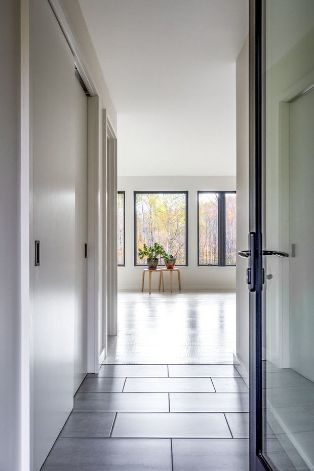 A bright, minimalist residential hallway leading into an open, unfurnished living area. The foreground has large-format grey floor tiles, white walls and built-in storage, while a dark-framed glazed door opens toward a room with pale timber flooring, expansive black-framed windows, and a small console table with potted plants. The space is clean, airy, and strongly lit by natural light.