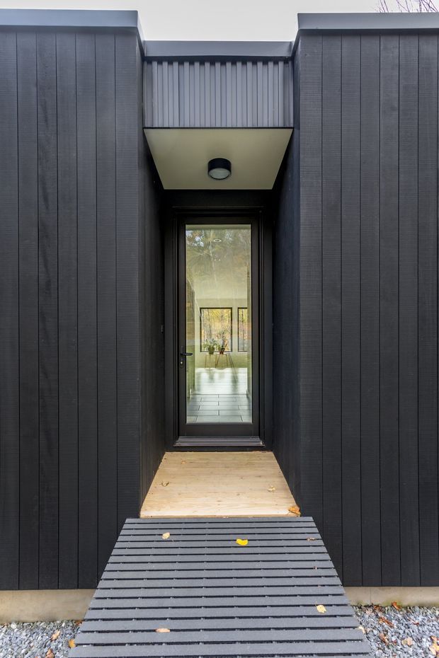 A narrow, minimalist exterior entry approach framed by dark vertical cladding. A black-framed glazed door sits beneath a projecting canopy with a recessed round light, with a timber landing and slatted dark ramp leading toward the entrance. Through the door, a bright interior with tiled flooring and further glazing is visible.