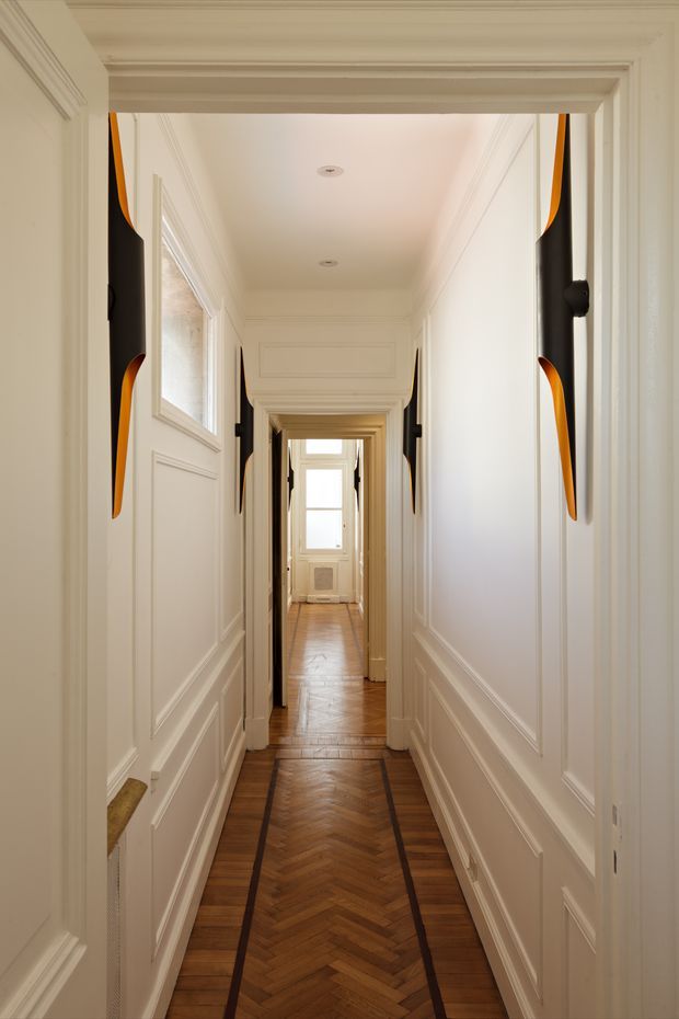 A long, narrow interior hallway with white paneled walls, ornate mouldings and cornices, recessed ceiling lights, and a polished herringbone timber floor bordered by dark inlay strips. Multiple doorways line the corridor, with daylight entering from a window at the far end. Black-and-orange sculptural wall-mounted accents add contrast.