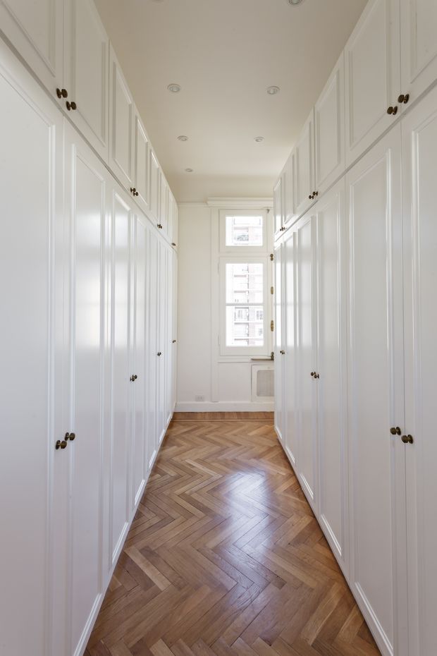 A narrow, bright walk-in wardrobe or dressing corridor lined on both sides with full-height white paneled built-in cabinetry and dark round knobs. The space has a light neutral ceiling with recessed downlights, a small multi-pane window at the end, and a warm mid-tone timber herringbone parquet floor.