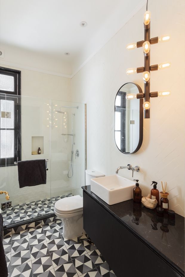A bright, contemporary bathroom with a walk-in glass shower, wall-mounted black vanity, white vessel basin, toilet, arched black-framed mirror, and a distinctive vertical brass-and-glass sconce. The room features bold geometric black, white, and grey patterned floor tiles, white textured wall tiles, and a black-framed window.