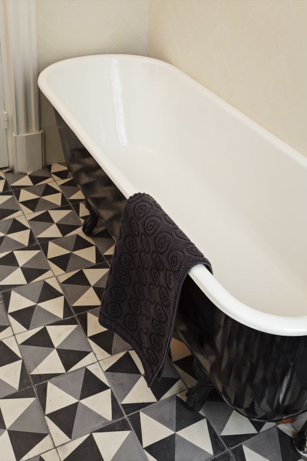 A close-up view of a freestanding white bathtub with a glossy black exterior and curved clawfoot-style base. The bathroom features striking black, white, and grey geometric floor tiles, pale patterned wall tiles, and a dark textured towel draped over the tub edge.