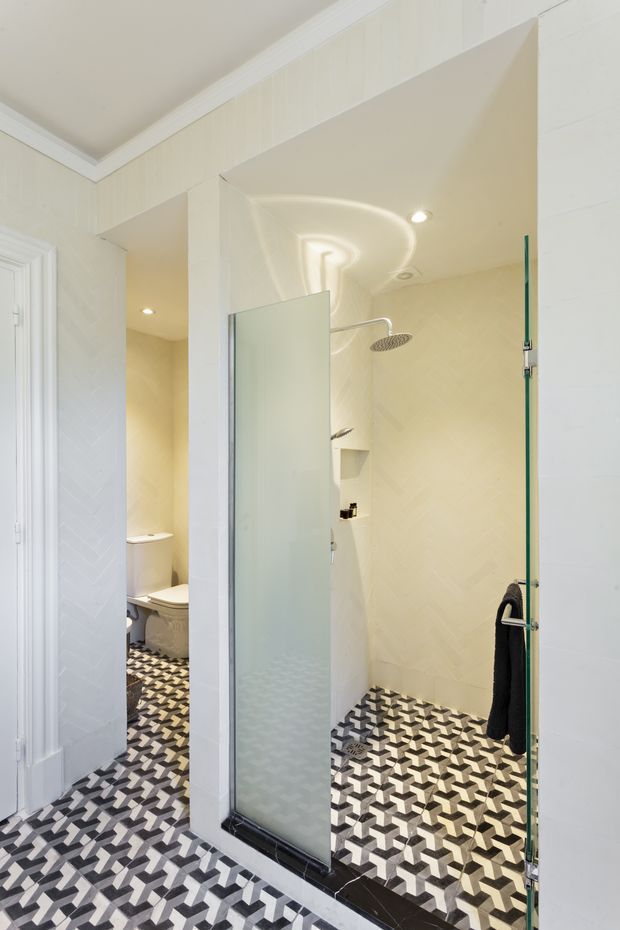 Bright contemporary bathroom featuring a walk-in glass shower with a frameless glass screen and large round rainfall showerhead. The space has pale cream geometric wall tiles, bold black, white and grey patterned floor tiles, white trim, recessed lighting and a partially visible toilet area.