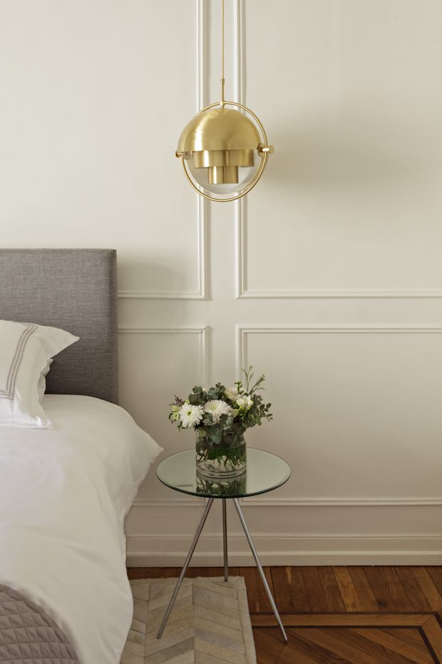 A refined bedroom vignette featuring a partially visible upholstered bed with white bedding, a small round glass-top side table, and a flower arrangement. The room has pale cream decorative wall panelling, timber parquet flooring, and a sculptural brass pendant light suspended above the table. The styling is elegant, calm, and traditionally detailed with contemporary furnishings.