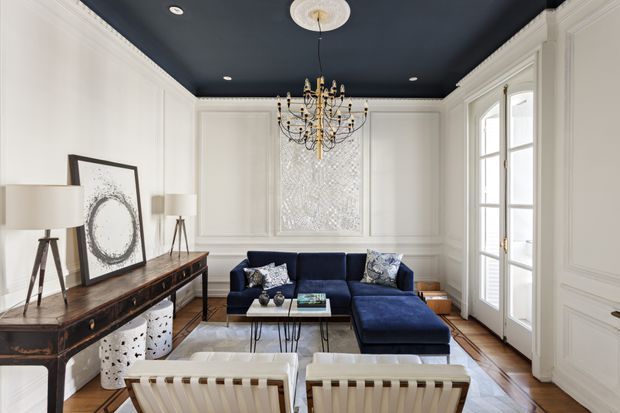 Elegant formal living room with white paneled walls and detailed crown moulding, a deep navy-blue ceiling, dark blue sectional sofa, dark wood console, framed artwork, and a statement crystal chandelier. Tall French doors and pale furnishings bring in natural light, while the blue-and-white palette creates a refined, high-contrast atmosphere.