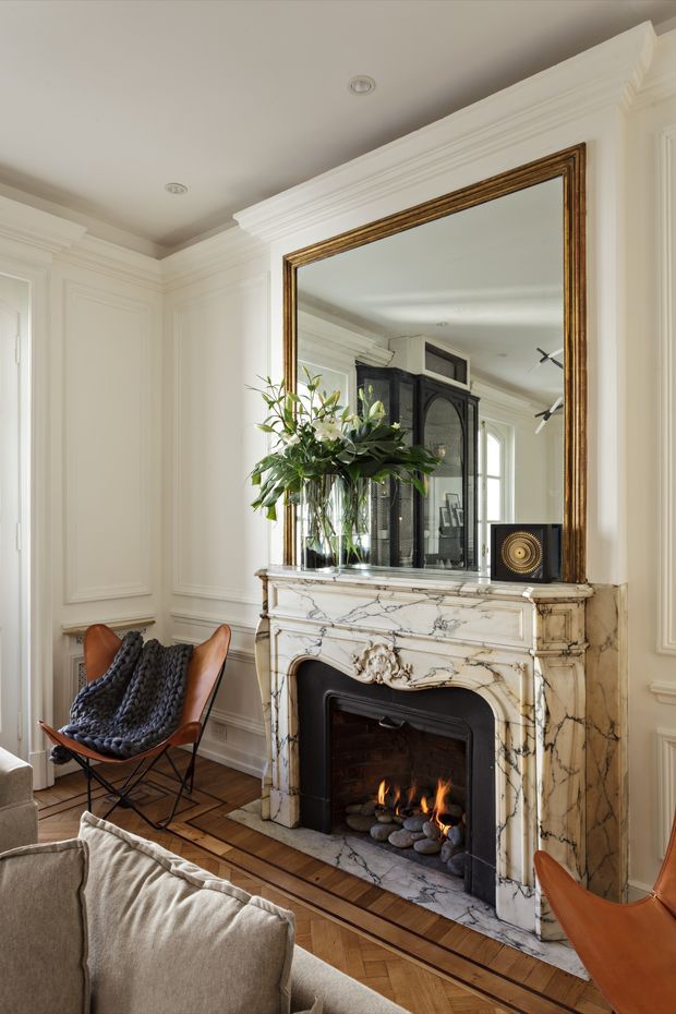 Elegant traditional living room featuring a carved marble fireplace with a lit fire, oversized gilded mirror, ornate wall mouldings, herringbone timber flooring, and leather lounge chairs. The room is styled with a restrained, refined palette of warm white, marble, timber, and dark accents.