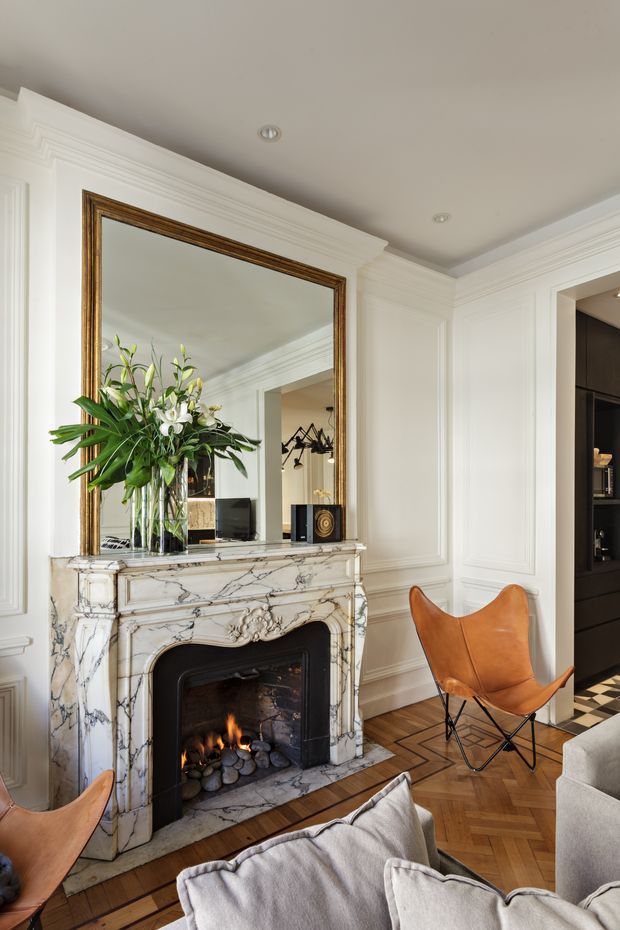 Elegant living room centred on an ornate veined marble fireplace with a lit fire, large framed mirror, decorative wall moulding, and a floral arrangement. The room includes a tan leather butterfly chair, pale upholstered seating, herringbone timber flooring, and a dark built-in cabinet visible through the opening at right. The interior has a refined, classic character with contemporary furnishings.