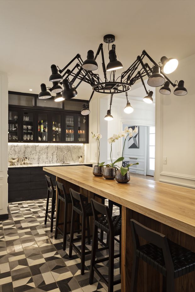 A sophisticated kitchen and entertaining space featuring a long light timber island with dark bar stools, black built-in cabinetry and a drinks display, a marble-look backsplash, and a dramatic cluster of articulated black pendant lamps. The room has decorative wall mouldings, orchids on the island, and a bold geometric black, white, and grey tiled floor.