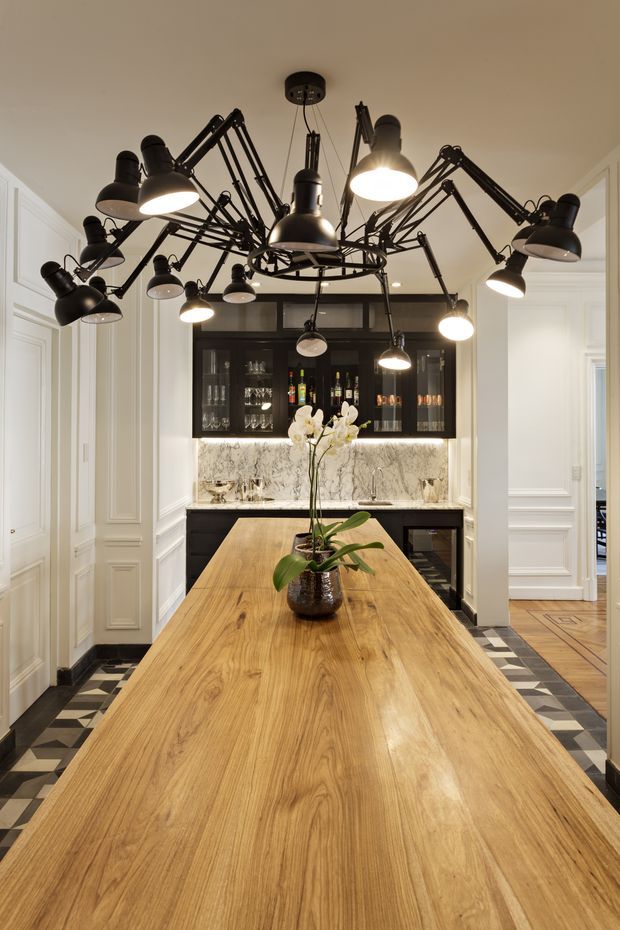 A refined kitchen and entertaining space viewed along a long timber island. The room features white decorative wall panelling, a dramatic black multi-arm articulated pendant light, black-framed glass cabinetry, a marble-look splashback, dark cabinetry, and a patterned black-and-white floor border. A large orchid arrangement sits on the island, with a glimpse of an adjacent hallway beyond.