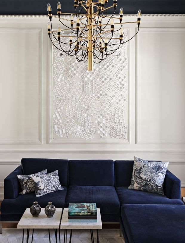 A refined living room featuring a deep navy sectional sofa with patterned monochrome cushions, two marble-topped coffee tables, and sculptural dark decorative vessels. The walls are finished with ornate white panel moulding and a textured white inset artwork, while a dramatic brass-and-black multi-arm chandelier hangs overhead. The overall composition is formal, layered, and high-contrast.