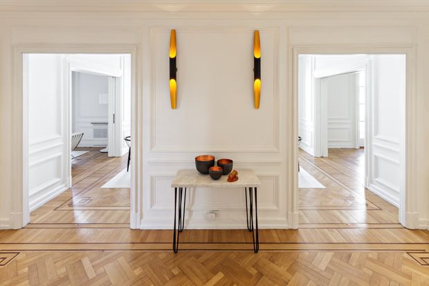 Elegant, bright residential hallway or entry gallery with ornate white wall panelling, two matching vertical wall sconces, and a slim console table. Wide openings on both sides reveal adjoining rooms, while the foreground features richly patterned parquet timber flooring with decorative border detailing. The space has a refined, symmetrical, classic character.