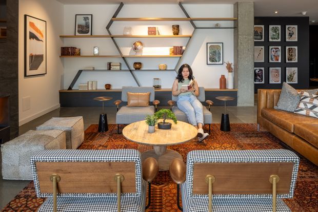 A warm, carefully styled living room with custom asymmetrical open shelving spanning the rear wall, displaying books, ceramics and framed artwork. Two patterned lounge chairs face a round timber coffee table, with a grey sofa, leather sectional, ottomans and layered patterned rugs completing the seating arrangement. The room features a textured stone or concrete-look column, timber shelving, neutral walls, and a mix of contemporary and mid-century-inspired furniture.