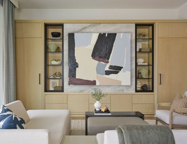 A refined living room with a full-width light timber built-in media and display wall, central abstract artwork, open shelving, and concealed cabinetry. The room includes a pale sectional sofa, upholstered armchairs, a dark rectangular coffee table, textured rug, and layered neutral furnishings. Soft daylight enters through a curtained window, creating a calm, contemporary atmosphere.