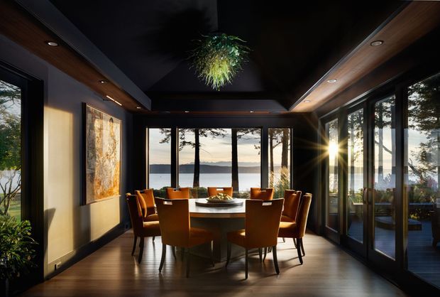 A sophisticated dining room with a large round dining table and upholstered chairs, framed by extensive floor-to-ceiling glazing overlooking a calm body of water and wooded landscape. The room features dark timber flooring, warm timber ceiling accents, dark painted ceiling panels, recessed lighting, extensive black-framed glazing, and a sculptural hanging plant fixture. A large artwork and indoor greenery add visual interest. The atmosphere is warm, refined, and dramatic, with strong indoor-outdoor connection.