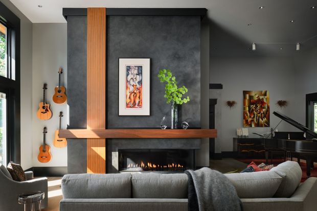 A sophisticated contemporary living room centred on a dark textured fireplace feature wall with a long timber mantel and vertical timber accent panel. A linear gas fire sits below framed artwork and a glass vase, while a large grey sectional sofa faces the fireplace. Guitars are displayed on the left wall, and a grand piano with artwork is visible to the right. Black-framed windows and recessed ceiling lighting complete the refined, moody interior.