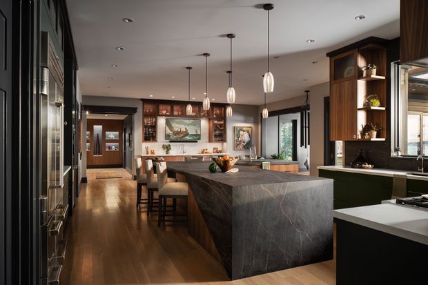 A spacious open-plan kitchen and dining area with a large dark stone-clad island, integrated seating, dark timber cabinetry, and a warm timber floor. Pendant lights hang above the island, while built-in shelving, artwork, display cabinetry, and a sink run along the perimeter. The room has a refined, high-end contemporary feel with strong contrast between dark joinery and lighter surfaces.