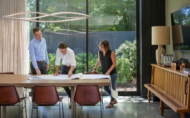 A bright contemporary dining and meeting space with a long timber table, leather dining chairs, and three people reviewing architectural plans. Floor-to-ceiling glazing opens the room to a leafy garden, while a sculptural linear pendant, dark vertical wall panelling, and a timber sideboard with a television define the interior.