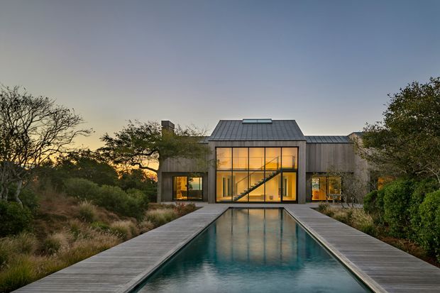 Contemporary two-storey residence viewed from the end of a long rectangular reflecting pool at dusk. The home features a dark standing-seam pitched roof, timber-clad wings, expansive floor-to-ceiling glazed walls, and a strongly symmetrical connection between the pool terrace and illuminated interior. Natural landscaping surrounds the property.