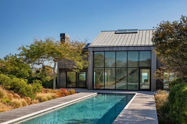 Contemporary dark-clad house viewed from the landscaped pool area, featuring a steep metal roof with skylight, expansive black-framed floor-to-ceiling glazing, and a long rectangular swimming pool bordered by timber decking. Mature planting surrounds the building.
