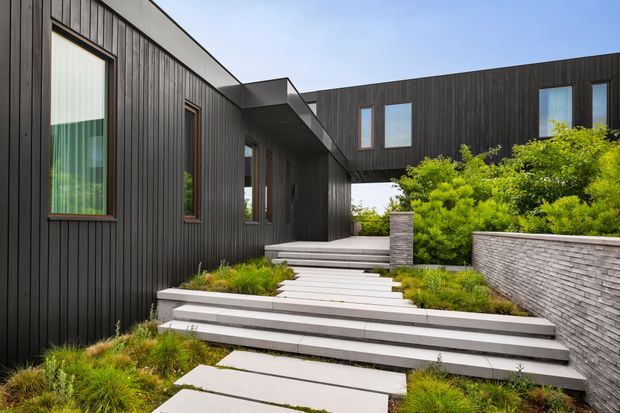 Modern exterior view of a dark-clad, multi-level residence arranged around a landscaped entry path. Broad concrete steps and floating rectangular pavers lead through ornamental grasses and shrubs toward recessed glazing and a covered entry. The building features vertical dark cladding, sharply projecting flat roof forms, narrow timber-toned window frames, and a textured grey masonry retaining wall.