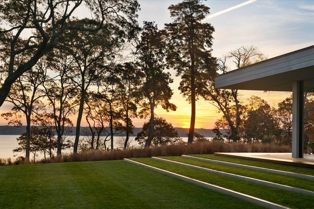 A modern flat-roofed pavilion or house extension sits at the edge of a manicured lawn, opening toward a calm body of water and a glowing sunset. Broad concrete steps with narrow illuminated or contrasting edges lead across the lawn toward the covered outdoor living area. The building has crisp horizontal roof lines, slender dark structural posts, and expansive open glazing or openings, framed by mature native-looking trees and long grasses.
