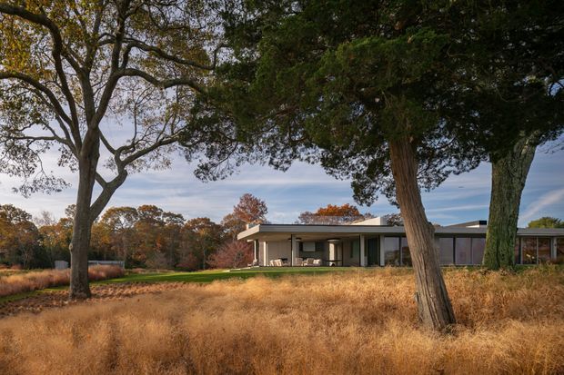 A low, single-storey modern residence set within a broad rural landscape. The house has a long, flat roofline, expansive glazing, pale rendered walls, dark vertical structural elements, and a covered outdoor terrace. Mature trees frame the view, with dry golden grass and autumn foliage surrounding the property.